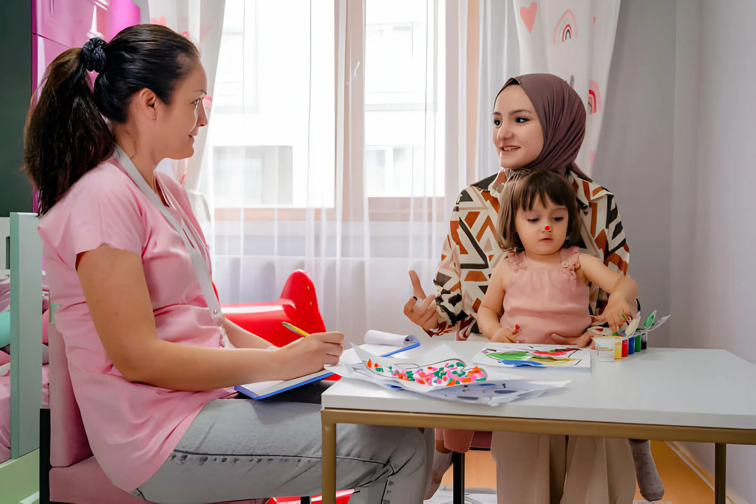 A healthcare professional takes notes during a consultation with a mother and her young child in a brightly lit pediatrician's office.