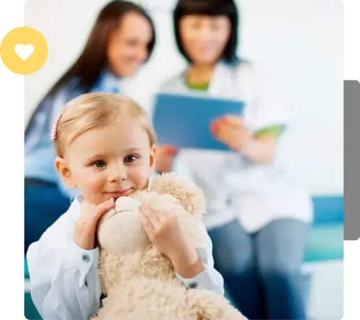 A young child clutching a teddy bear looks towards the camera with a gentle smile, while in the background, two healthcare professionals are in discussion, slightly out of focus.